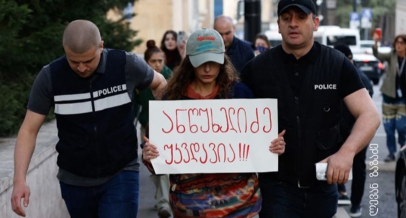 Полицейские ведут активистку, держащую плакат с надписью: "Анцухелидзе бессмертен!". Фото: Леван Зазадзе / Pirveli https://tvpirveli.ge/ka/siaxleebi/sazogadoeba/103394-aqtivisti-plakatit-antsukhelidze-ukvdavia-5000-larit-daajarimes