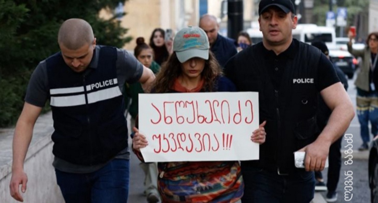 Полицейские ведут активистку, держащую плакат с надписью: "Анцухелидзе бессмертен!". Фото: Леван Зазадзе / Pirveli https://tvpirveli.ge/ka/siaxleebi/sazogadoeba/103394-aqtivisti-plakatit-antsukhelidze-ukvdavia-5000-larit-daajarimes